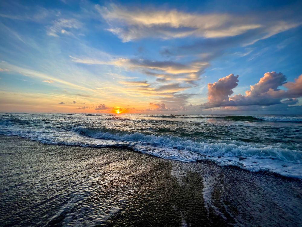 Orange sun rises over the ocean as waves recede from the shore leaving dappled water on the dark sand. Orange and grey clouds drift on the horizon. 
