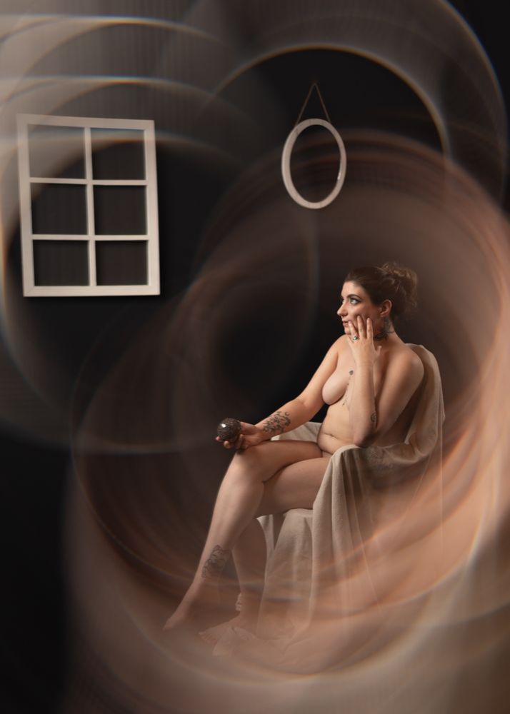 A color photo of a nude woman sat on a chair covered in a canvas drape. She is holding a glass bauble and touching her face pensively. She is surrounded by ethereal auras.