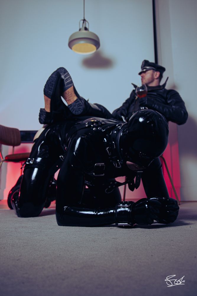 Man in full leather, drone knelt in full rubber in front. Boots on drone's back.