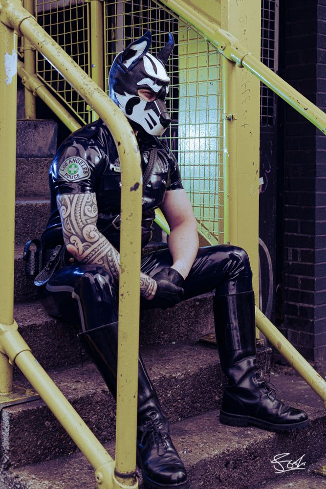 Rubber uniform clad human pup wearing rubber uniform and tall patrol boots, sitting on car park steps.