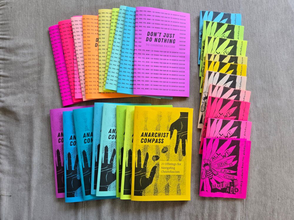 Multiple copies of three zines laid out in rainbow colors on a beige couch. The zines are "don't just do nothing to counter fascism, "anarchist compass: 29 offerings for navigating chrisofascism," and "all we have is each other: mutual aid"