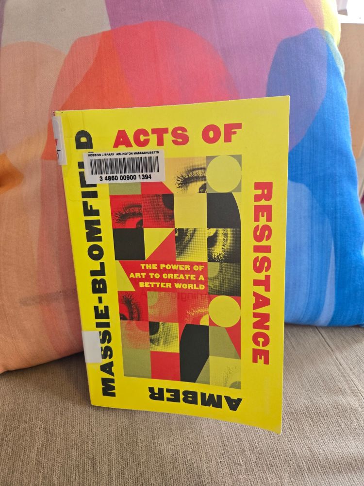 The book Acts of Resistance: The Power of Art to Create a Better World by Amber Massie-Blomfield, leaning against a colorful abstract throw pillow. The book cover is yellow, red, and black, and features a collage of squares with photos of eyes and geometric shapes