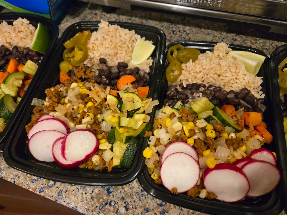 Plastic takeout containers full of beans, rice, zucchini, carrots, onions, corn, seitan chorizo, lime wedges, radish slices, and pickled jalapenos