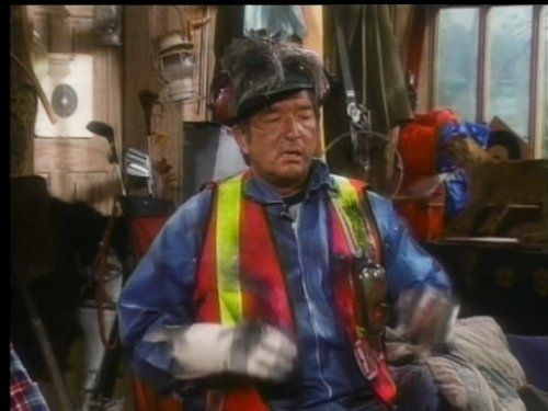 Graham Greene wearing a helmet and orange hi-vis vest covered in burn marks from the Red Green Show