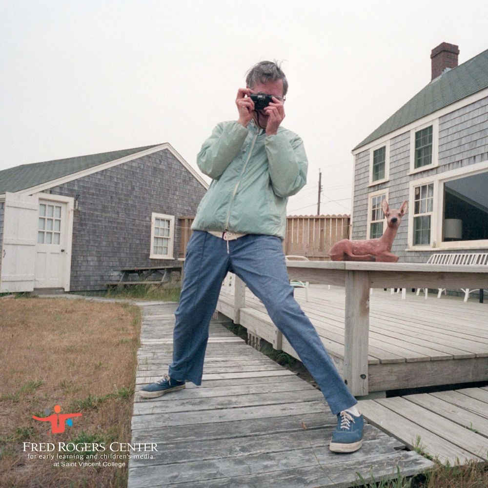Fred Rogers standing in a wide stance on a walkway near a porch, aiming a camera at the person taking the photo. Pastel green windbreaker, canvas sneakers, sculpture of a baby deer, dead grass, overall New England goodness