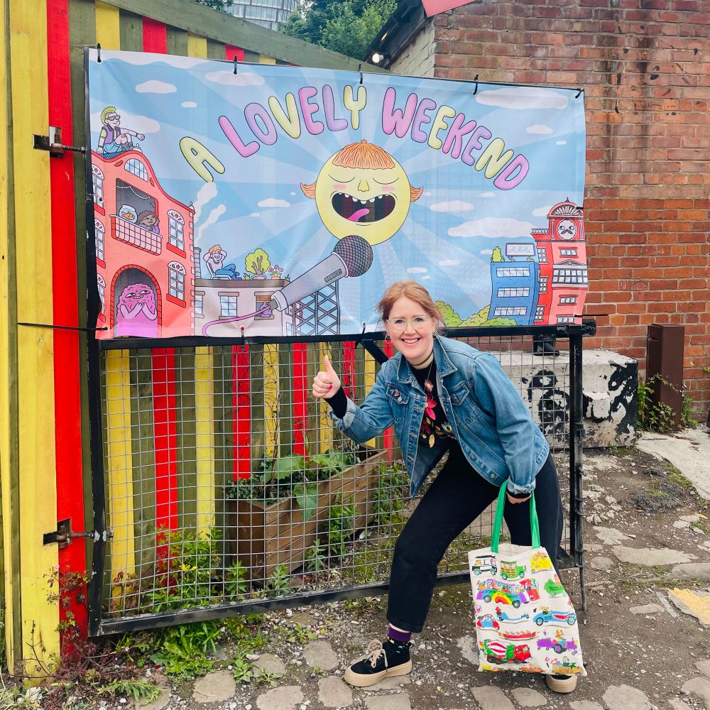 A woman (me) standing outside, in front of a venue with a big illustrated banner hanging up. The banner is for A Lovely Weekend comedy festival and is covered in summery cartoon illustrations of buildings and weird little characters.