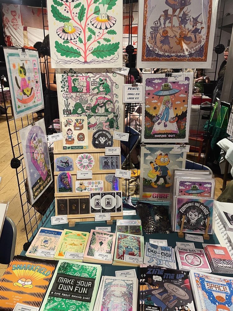 My stall at zine fest! Loads of zines, prints, pins, stickers and bookmarks are displayed on a blue tablecloth. 
