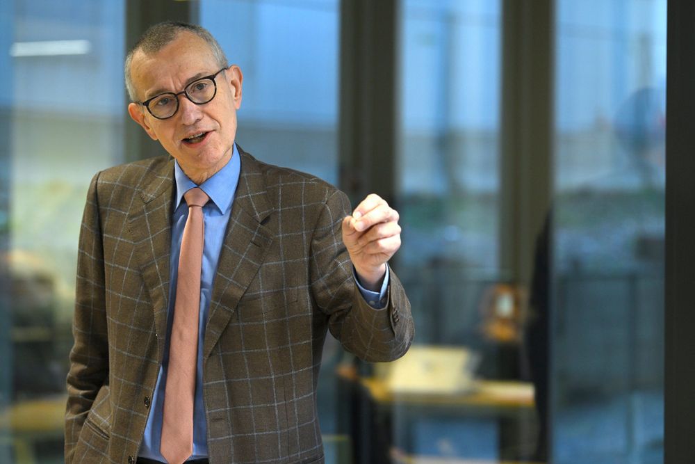 Belgian minister Franck Vanden Broecke. A distinguished man in a brown checkered suit, wearing a blue shirt and pink tie. One hand in his right pocket and gesturing with his left hand. 