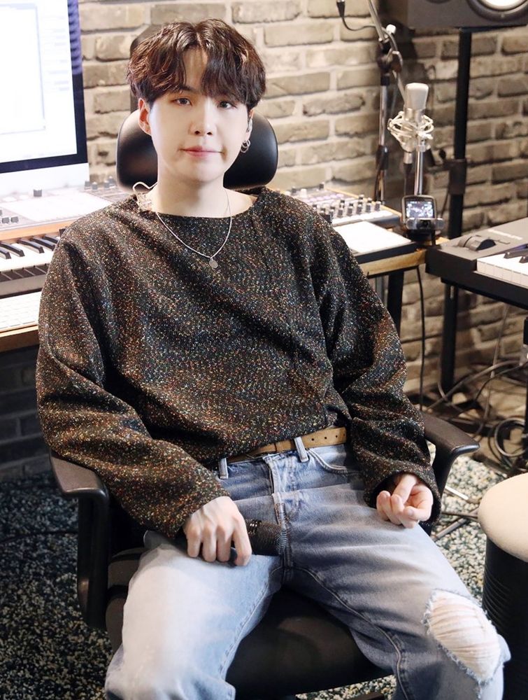 a cute dorky yoongi in a dark sweater and light jeans, on a chair, legs spread, in his studio, with his signature :] expression 