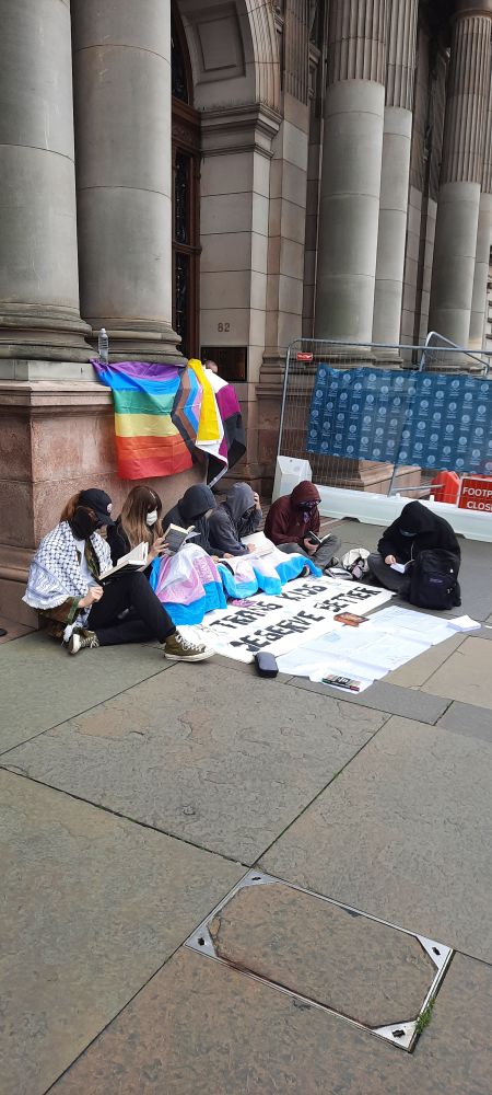 Photo of young people reading books outside the pillars of a large sandstone building. They have queer flags and a banner that reads trans kids deserve better.