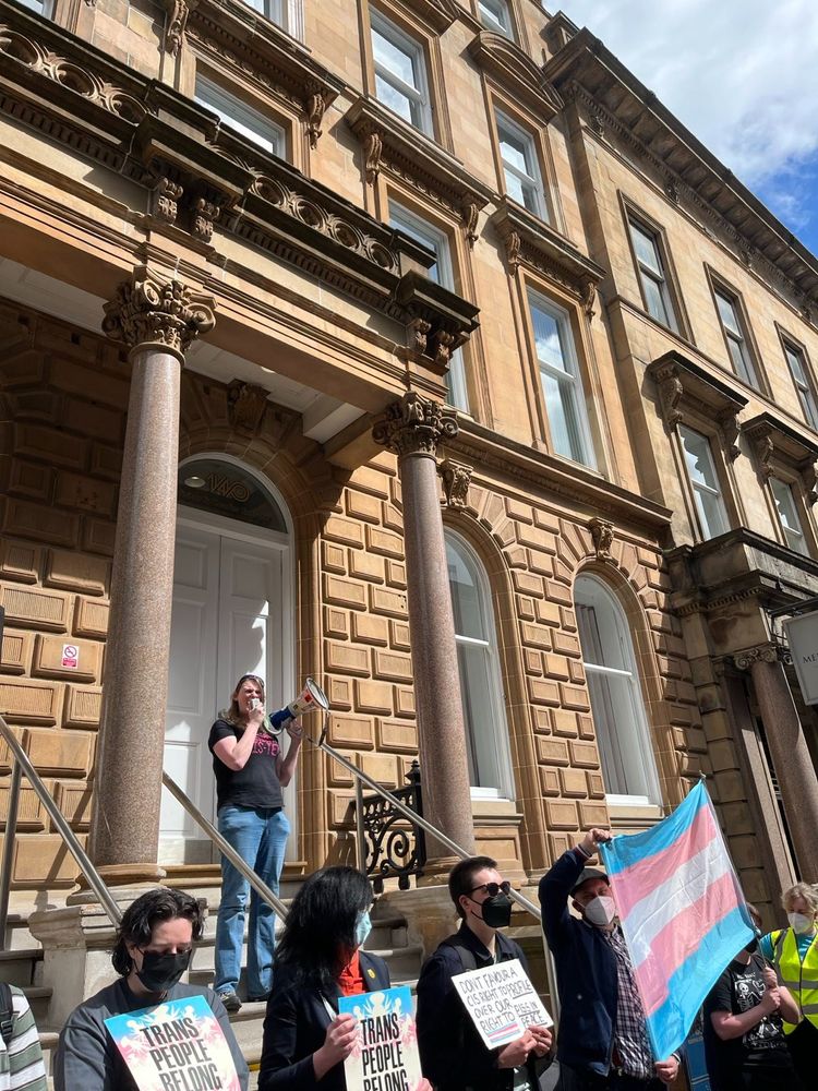 Photo of people protesting outside a Victorian building. A woman wearing jeans and t-shirts stands on steps speaking into a megaphone. Two people hold a trans flag, and others hold placards that read: trans people belong here.