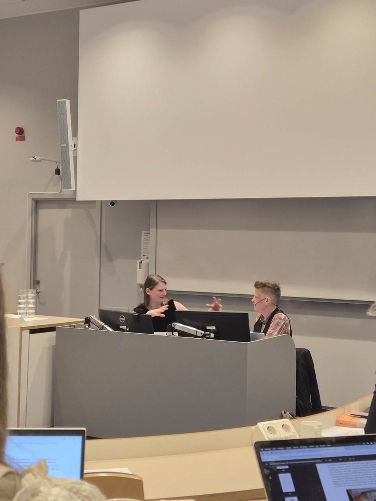 Photo of two people talking, seated behind a desk at the front of a lecture theatre. One is gesturing.