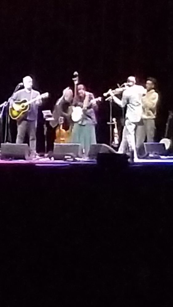Slightly fuzzy picture of an African American woman bent enthusiastically over a banjo, with a stylish dude in a white suit on the fiddle, white guy on guitar, and two backup players behind them.