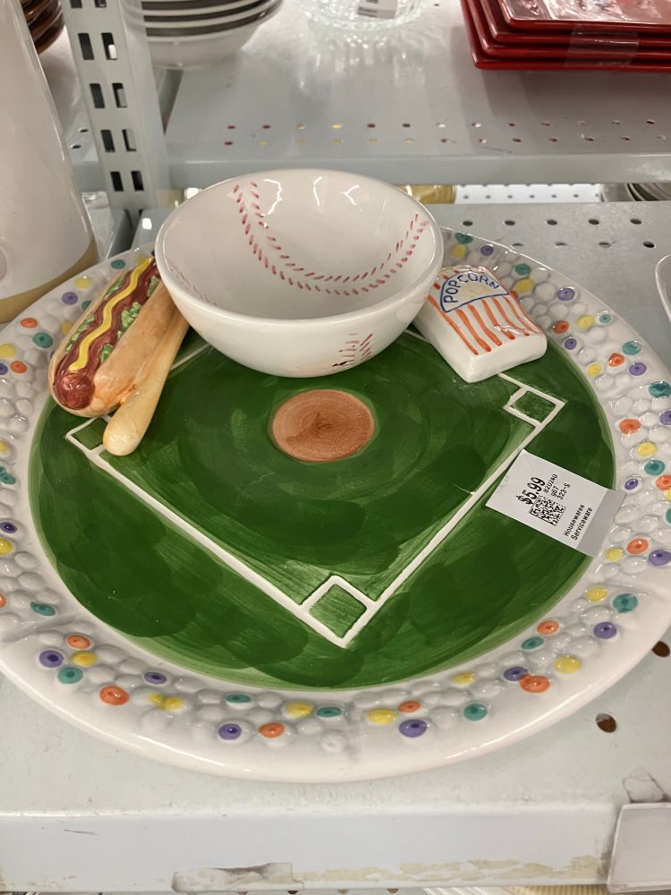 A ceramic dish painted like a baseball stadium with an attached bowl that is a half baseball. It also has a bag of popcorn, a hotdog, and a tiny baseball bat also ceramic 