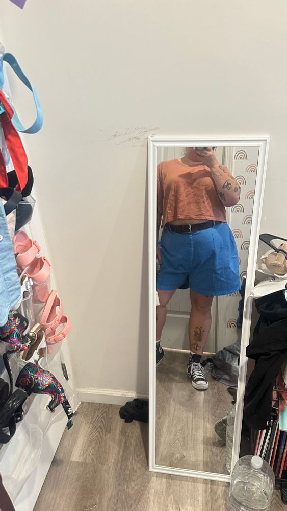 A photo of Katelyn in blue shorts and an orange t-shirt. They are also wearing converse and a black belt. 