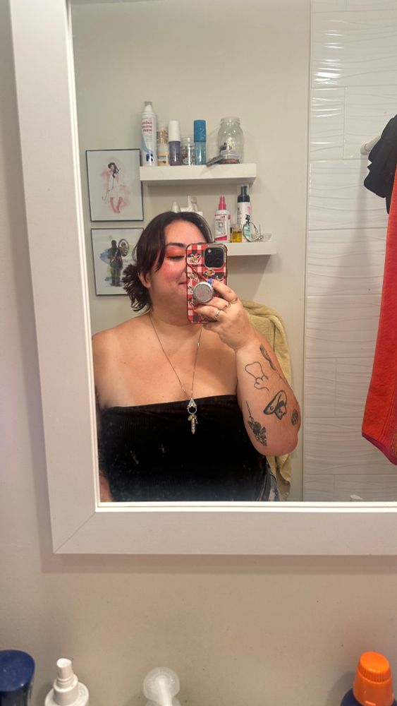 Katelyn taking a selfie in a mirror. They are wearing black tube top and a silver necklace. 