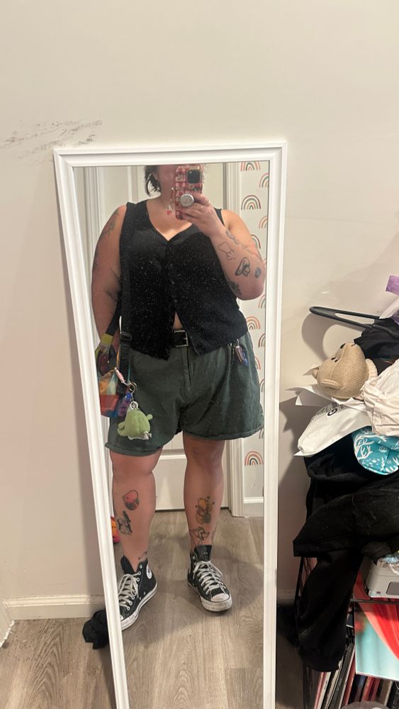 A photo of Katelyn wearing green shorts and a black tank top. They are also wearing converse and a purse. 