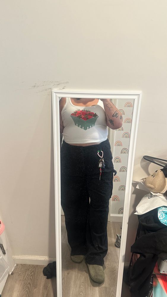 A photo of Katelyn wearing black pants and a tank top with strawberries on them. They are also wearing green Birkenstocks. 