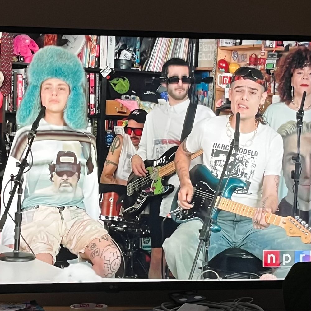 Foto do vídeo do Tiny Desk com Ca7riel & Paco Amoroso

Em primeiro plano, na esquerda, uma pessoa branca com um daqueles chapéus de inverno russos, cor verde-água, e, na direita, uma pessoa branca segurando uma guitarra e a camiseta escrito NARCOMODELO

A impressão é de que todos os integrantes são jovens e fluidos, e a sensação é de que são muito deslocados e desconstruídos 

Ao fundo, um homem branco, o baixista, usa um óculos de sol com outros dois óculos de sol grudados na lateral do que está no rosto dele, um de cada lado