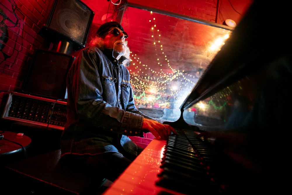 A man sits at a piano, surrounded by red light, as he leans back and plays. His hands clearly do not need his eyes for that. The piano keys stretch forward into the frame.