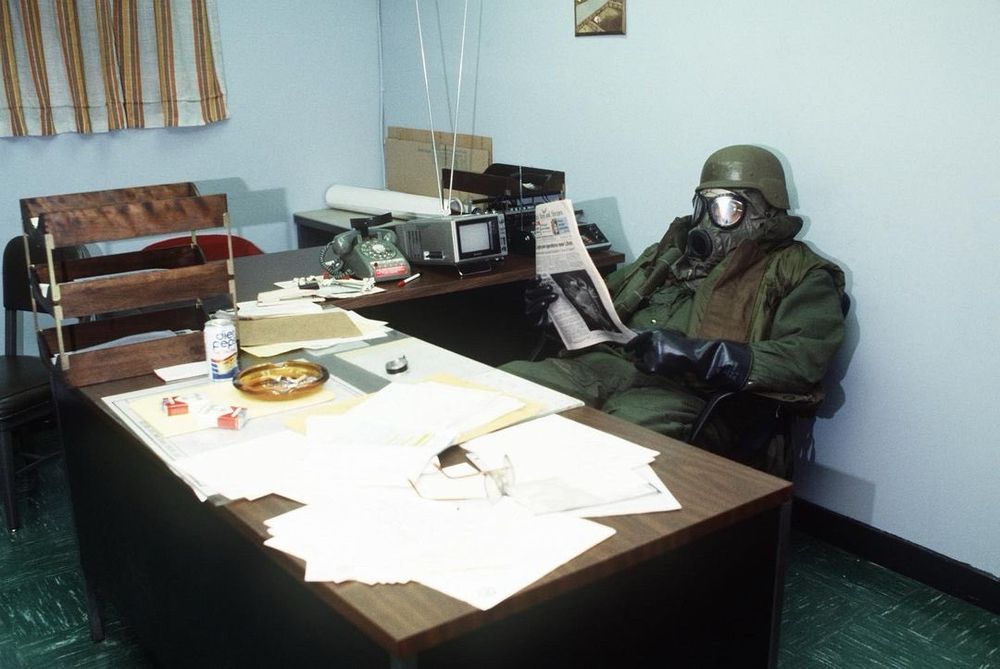 Someone in full 1980's/1990's military CBRN gear sitting at a desk scattered with papers, an ashtray, and a couple boxes of cigarettes. They're in an old, drab office, sitting back and reading a newspaper.