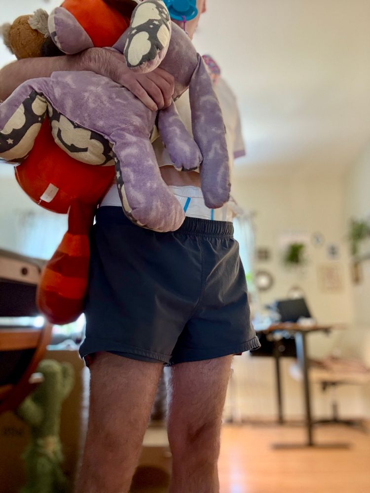 An adult is standing and proudly holding two stuffed animals (Jupiter, the rabbit; Mylo, the red panda). Their white diaper is visible poking out between the top of their shorts and the bottom of their crop top shirt. 