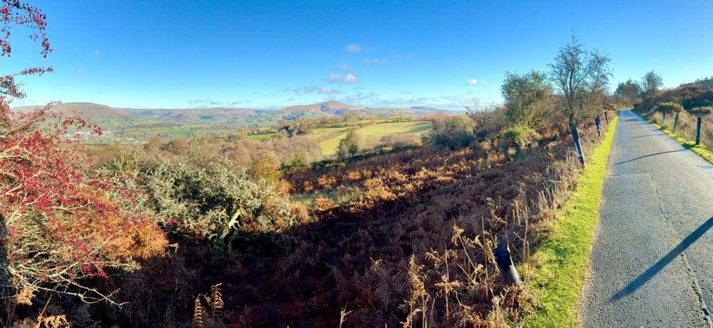 A panoramic of Hafod Road to the right, with far-reaching views over bracken, fields and Hawthorn trees stretching out to the left. The Sugarloaf mountain can just be seen on the horizon. Blue skies, long shadows and a warm light make for a beautiful winter’s day.