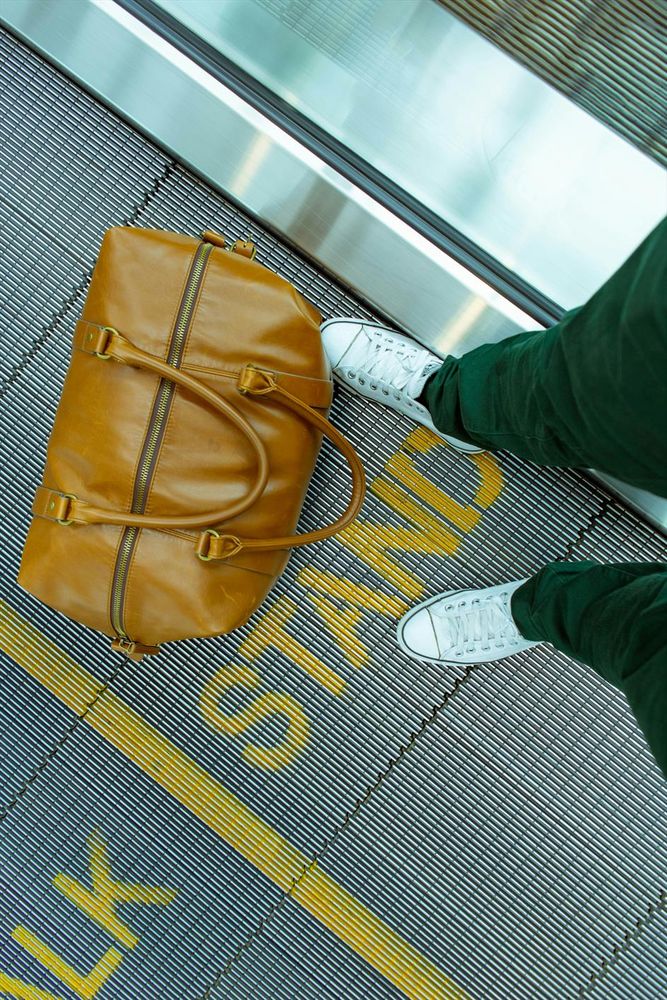 Photograph of a person stranding on a moving walkway in an airport beside a brown leather handbag. The word "stand" is written in yellow at their feet. By White Field Photo, under a free Unsplash license: https://bit.ly/3XNjLyr.