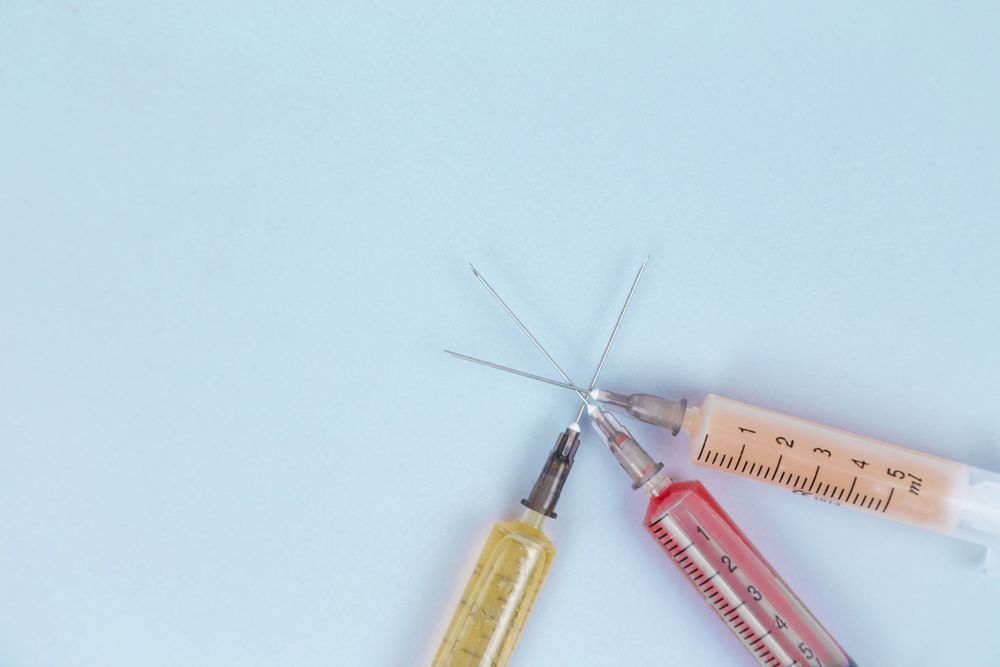 Photograph shows syringes with different medicines in yellow, red, orange and transparent colors that lie on a blue background. By Diana Polekhina under a free Unsplash license: https://bit.ly/4i2pjhj.