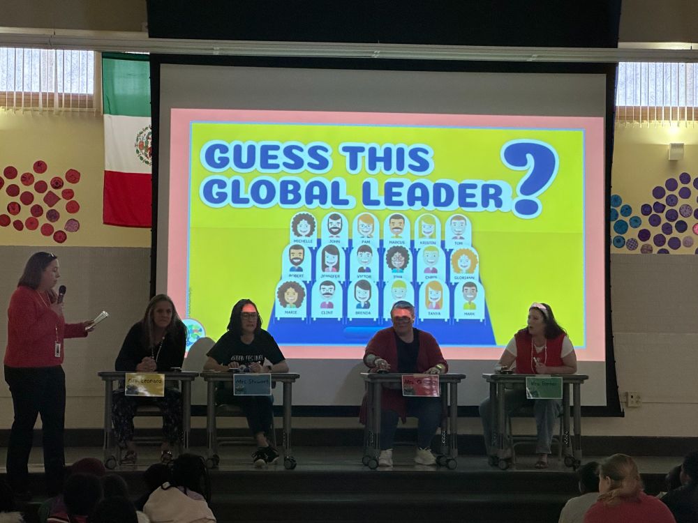 A group of individuals seated at tables in a school auditorium, participating in a quiz titled "Guess This Global Leader," displayed on a large screen behind them. Flags of different countries adorn the sides of the screen.