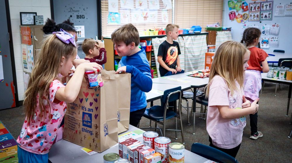 Children participating in a classroom activity, packing items into a bag and organizing canned goods on a table.