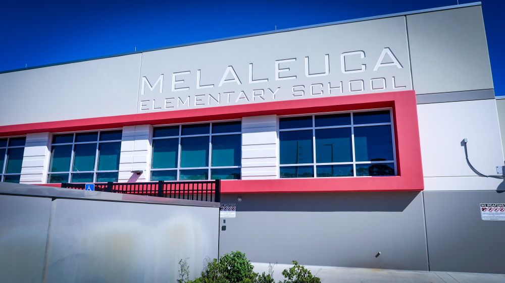 Exterior view of Melaleuca Elementary School with prominent signage, featuring a modern facade with red accents and multiple windows under a clear blue sky.
