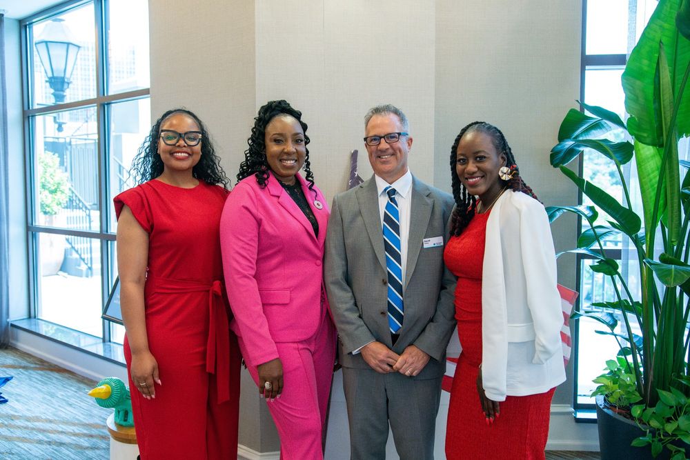 Four professionals posing for a photo at an indoor event. Two are dressed in red suits, one in a pink suit, and one in a grey suit. The setting includes a spacious room with large windows and a decorative plant in the background.
