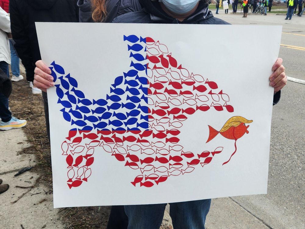 A school of tiny red, white and blue fish work together to form a giant fish that is shown eating an orange fish with Trump's distinctive comb over