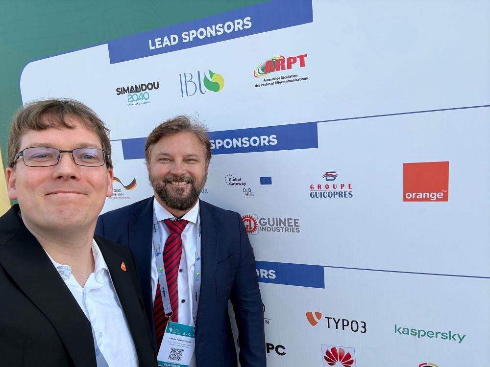 Two men in business attire smile for a selfie in front of a sponsor board at an event, featuring logos from various companies and organizations, including TYPO3.