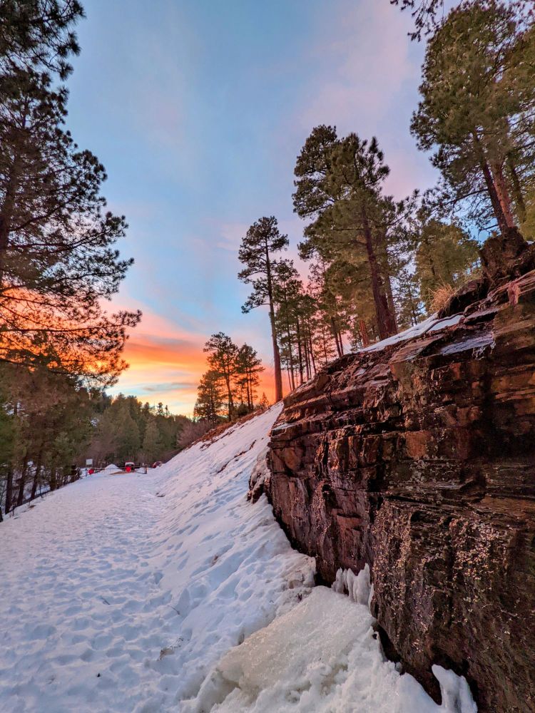 A photo of a mountain road covered in snow with a rock wall on the right side and tall conifer trees on both side of the road. A bright orange sunset is in the background of the photo. Taken on Mt. Lemmon in the Santa Catalina Mountains above the city of Tucson, Arizona.
