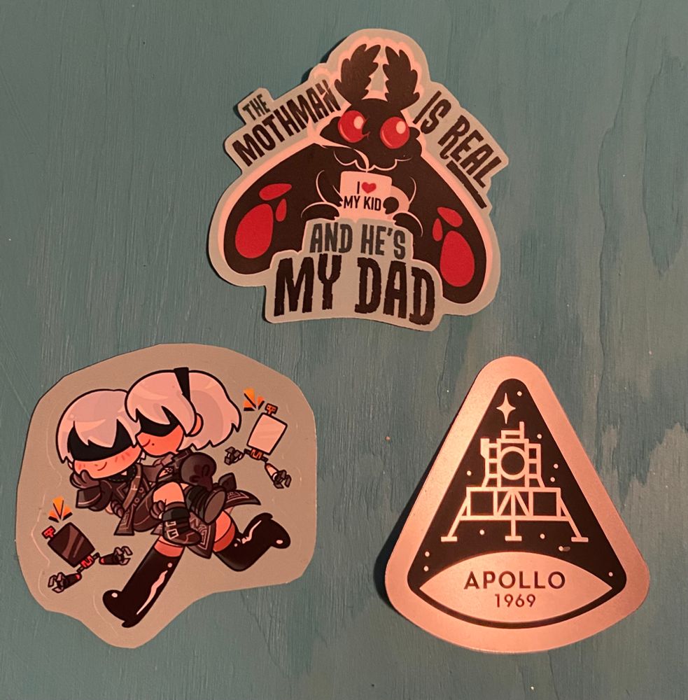 three stickers in a triangle
top sticker: Mothman holding a mug that says ‘I HEART my kid’, the text on the sticker says ‘The Mothman is real and he’s my dad’
bottom left sticker - 2B and 9S from Nier automata, 2B is princess carrying 9S and their pods are flying with them
bottom right sticker - a silver and black cone with the Apollo lunar lander that says Apollo 1969