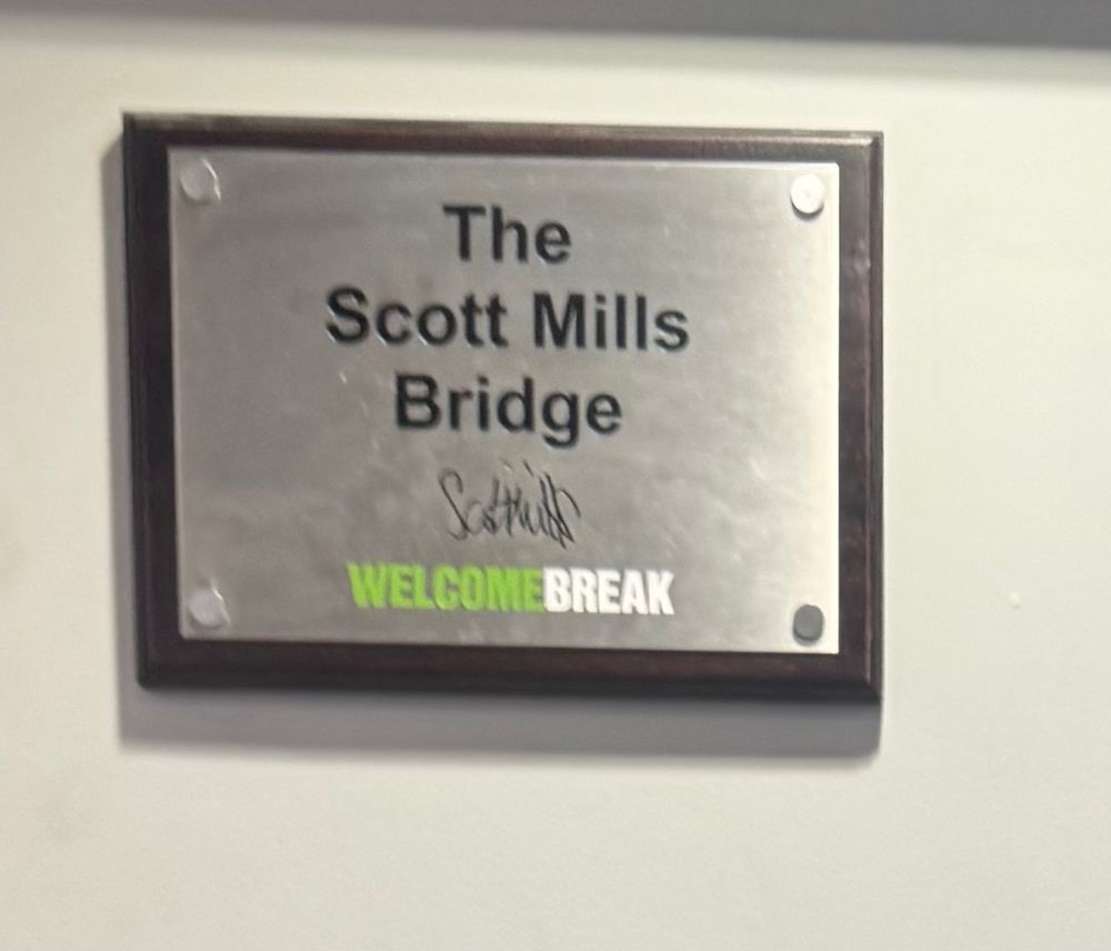 The sign for bridge named after Scott Mills at a Welcome Break services. It is one of those bridges that connects one half of the services with the other on the other side of the road. The sign also bears the signature of the man himself.