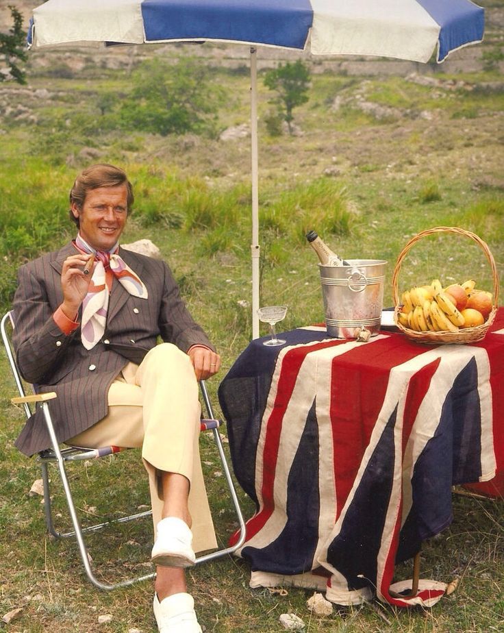 Roger Moore looking dapper and camp sitting next to a table covered in the union flag and bearing fruit and chilled champagne