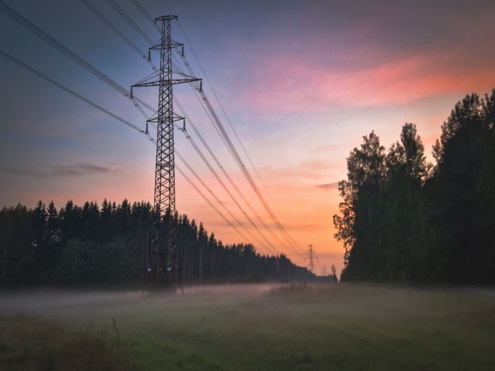 Sumuinen pelto auringon laskun kajossa jonka molemmin puolin puita, keskellä kulkee voimalinja 