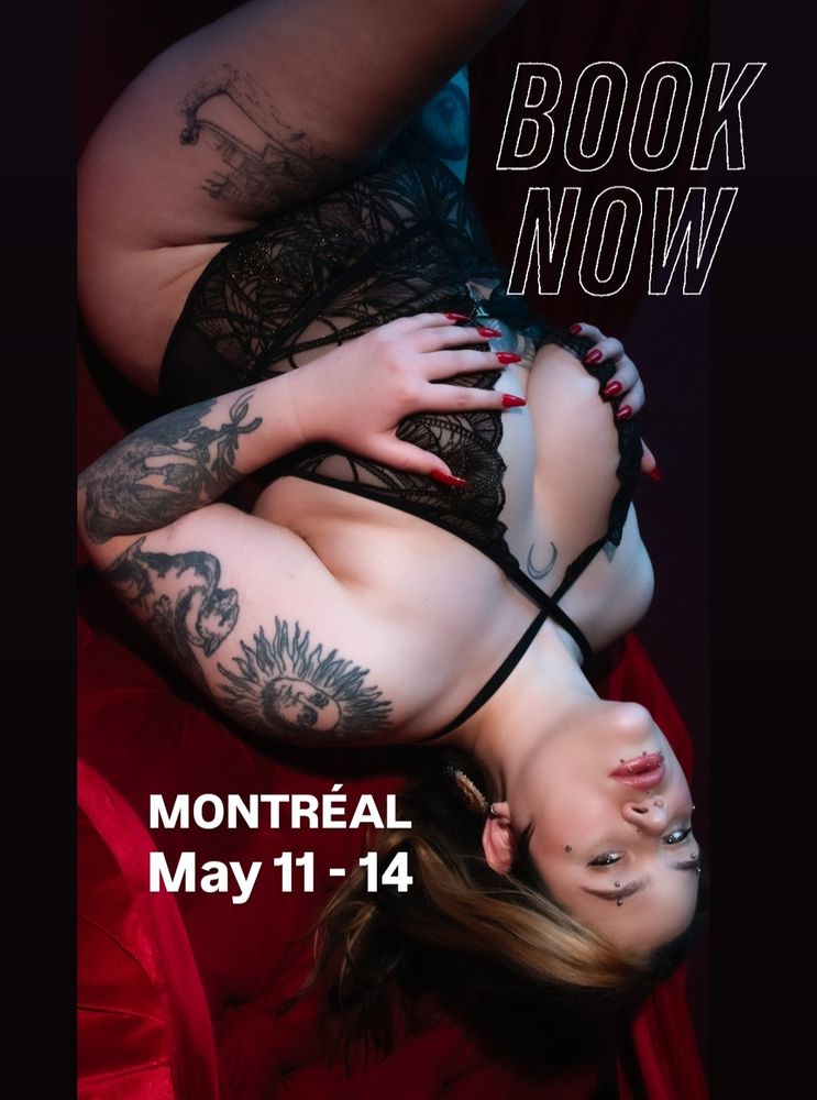Emilia laying on her back. She's looking into the camera. 
Wearing a black low cut bodysuit. 
Her hands cup her breasts.
One leg Is raised over the other.