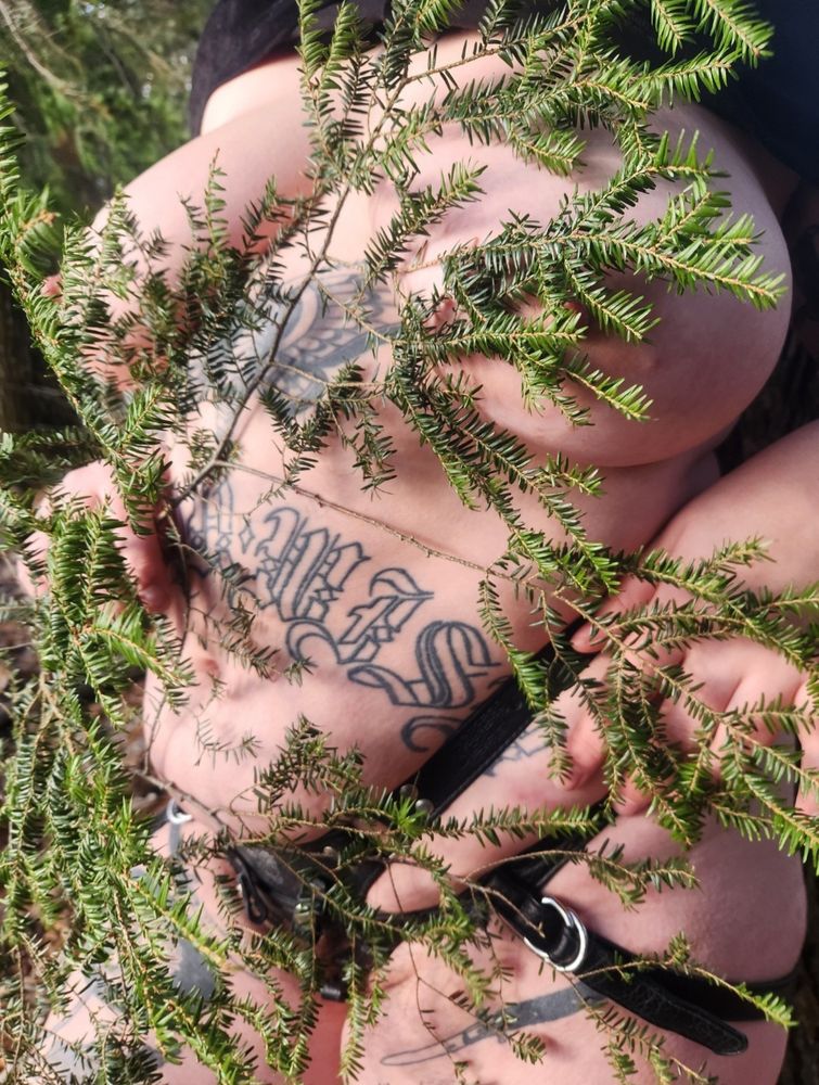 Emilia outside in the woods, a curvy tattooed white femme. Wearing a leather strap and holding foliage (eastern hemlock I think) in front of her body.