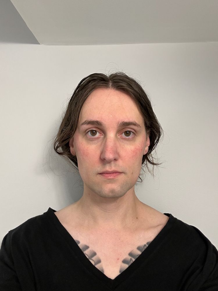 A selfie taken on October 1, 2024 of Jaen, a white trans femme with long brown hair that's parted in the middle and tied back. She's wearing a black v-neck shirt, has a neutral expression, and doesn't have any makeup on.