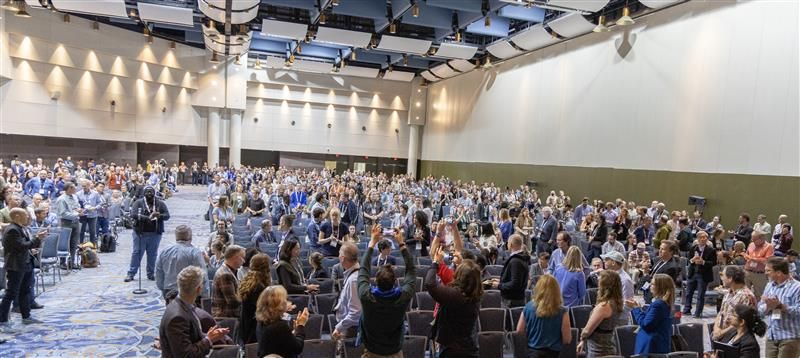 In a large conference room full of people, the audience doesn't fit entirely in the frame of a photo. Nearly everyone pictured is standing.