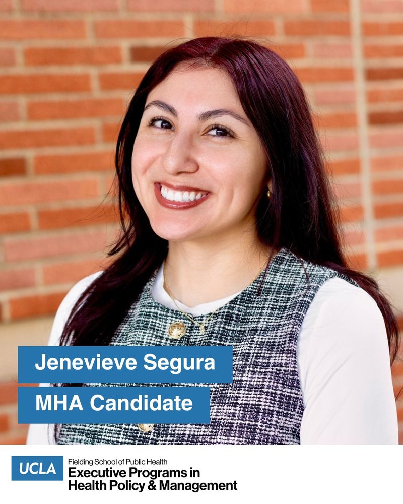 UCLA MHA Candidate Jenevieve Segura stands outside with a red brick wall behind her, on the UCLA campus. She has long dark hair and she wears a white long-sleeve top under a black and white tweed vest with gold buttons.