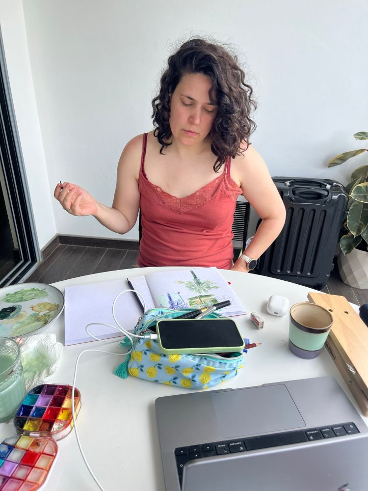 Photo of saur sitting at a table with a sketchbook, gouache and other clutter in it. She’s lifting a brush in one hand and her other hand is pressed into her hip while looking at the sketchbook page
