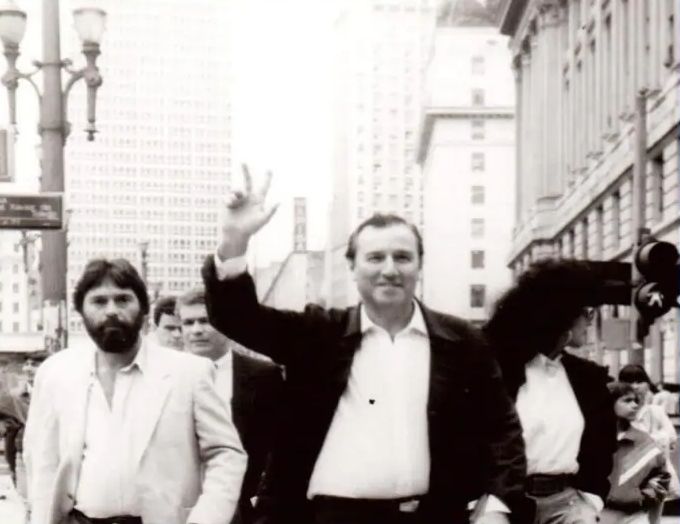 Foto em preto e branco. Eymael, mais novo, caminha pela rua, com um braço erguido. Veste camisa branca e paletó preto. Ele sorri. Há mais pessoas na foto. Há prédios e um poste com uma placa.