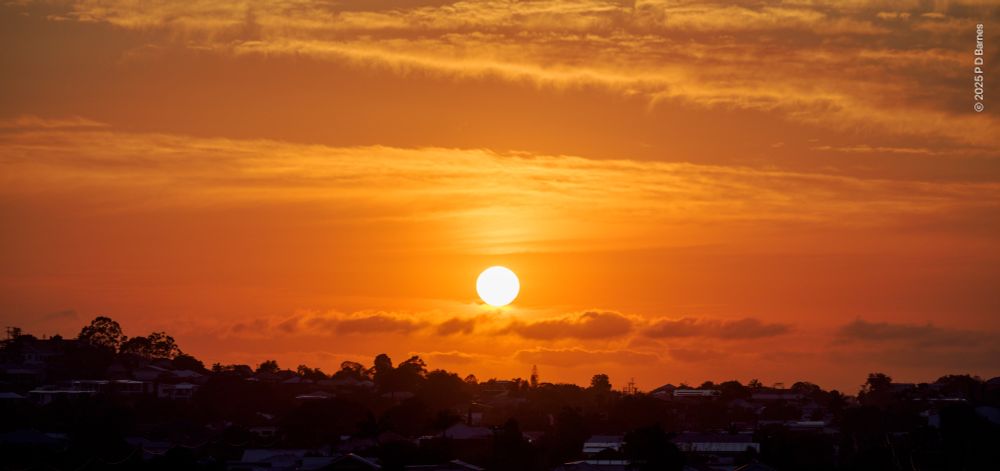 A blazing yellow sun in a golden sky. Several cloud systems are all backlit or transilluminated by the bright sun. Below, a suburban ridgeline and hillside is just visible against the bright sky