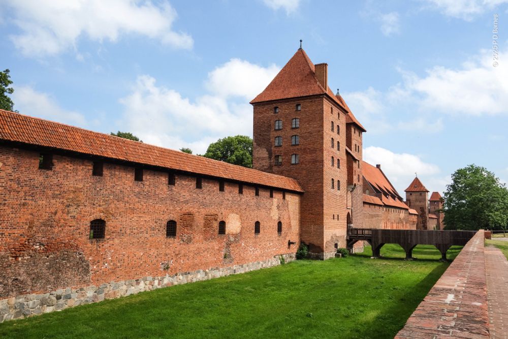 View down the length of part of one outer wall of Malbork Castle. It's built entirely of bricks and roofed with red terracotta tiles. The wall is about three stories high, punctuated with small window opening, and interrupted periodically by much higher six or seven storey towers. Immediately outside the wall is a wide grassed area like a moat, but dry. The nearest tower is paired and forms the entrance for a wooden footbridge.