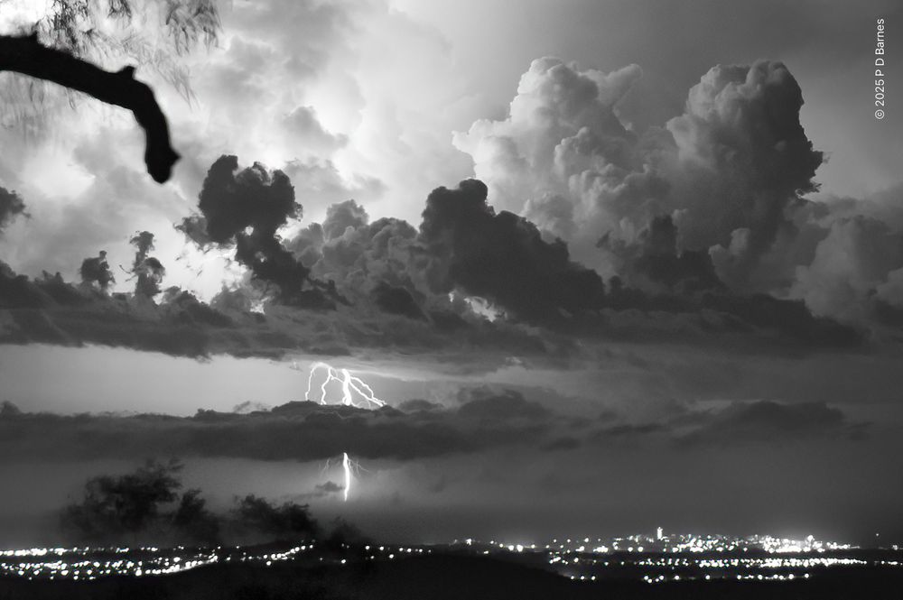 A large lightning bolt striking out to sea at night, seen from a high vantage point looking out over the coastal plains and lights of Caloundra. The lightning has lit up the storm's cloud banks dramatically.